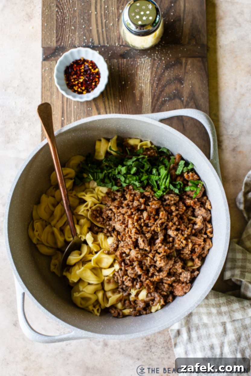 Overhead photo of a large pot filled with cooked Italian sausage, tortellini and basil