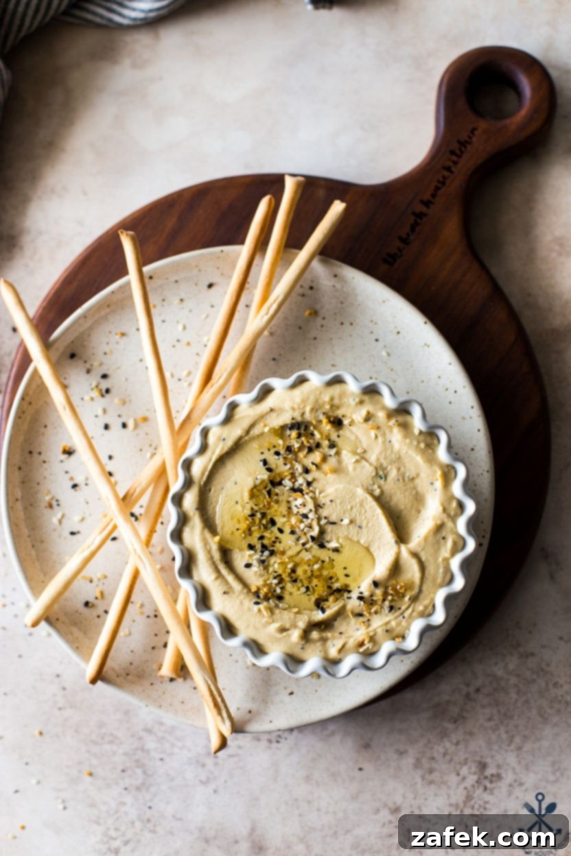 Hummus Unpacked 2 Overhead photo of a small dish of hummus on a plate with breadsticks, topped with everything bagel seasoning and a drizzle of olive oil.