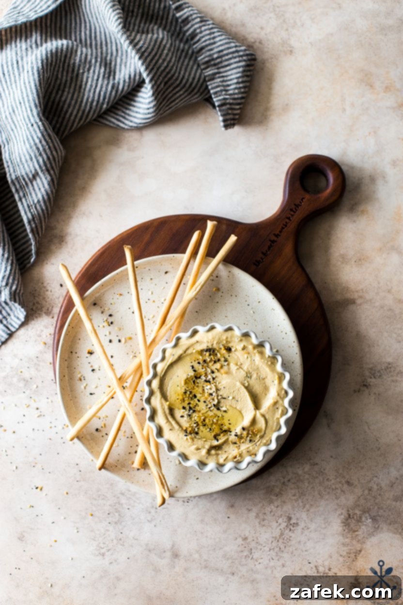 Hummus Unpacked 3 Overhead photo of a dish of hummus on a plate with thin breadsticks, garnished with olive oil and everything bagel seasoning.
