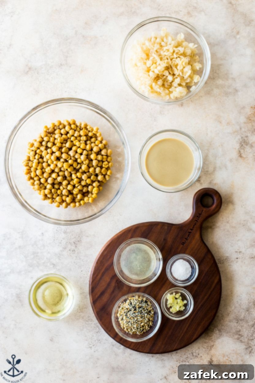 Hummus Unpacked 4 Overhead photo displaying all the fresh ingredients needed for making everything hummus: canned chickpeas, tahini, olive oil, lemon, garlic, sea salt, everything bagel seasoning, and red pepper flakes.
