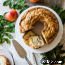 Old Fashioned Apple Spice Cake 13 Overhead photo of a baked Jewish Apple Cake on a white cake plate surrounded by fresh apples and green leaves.