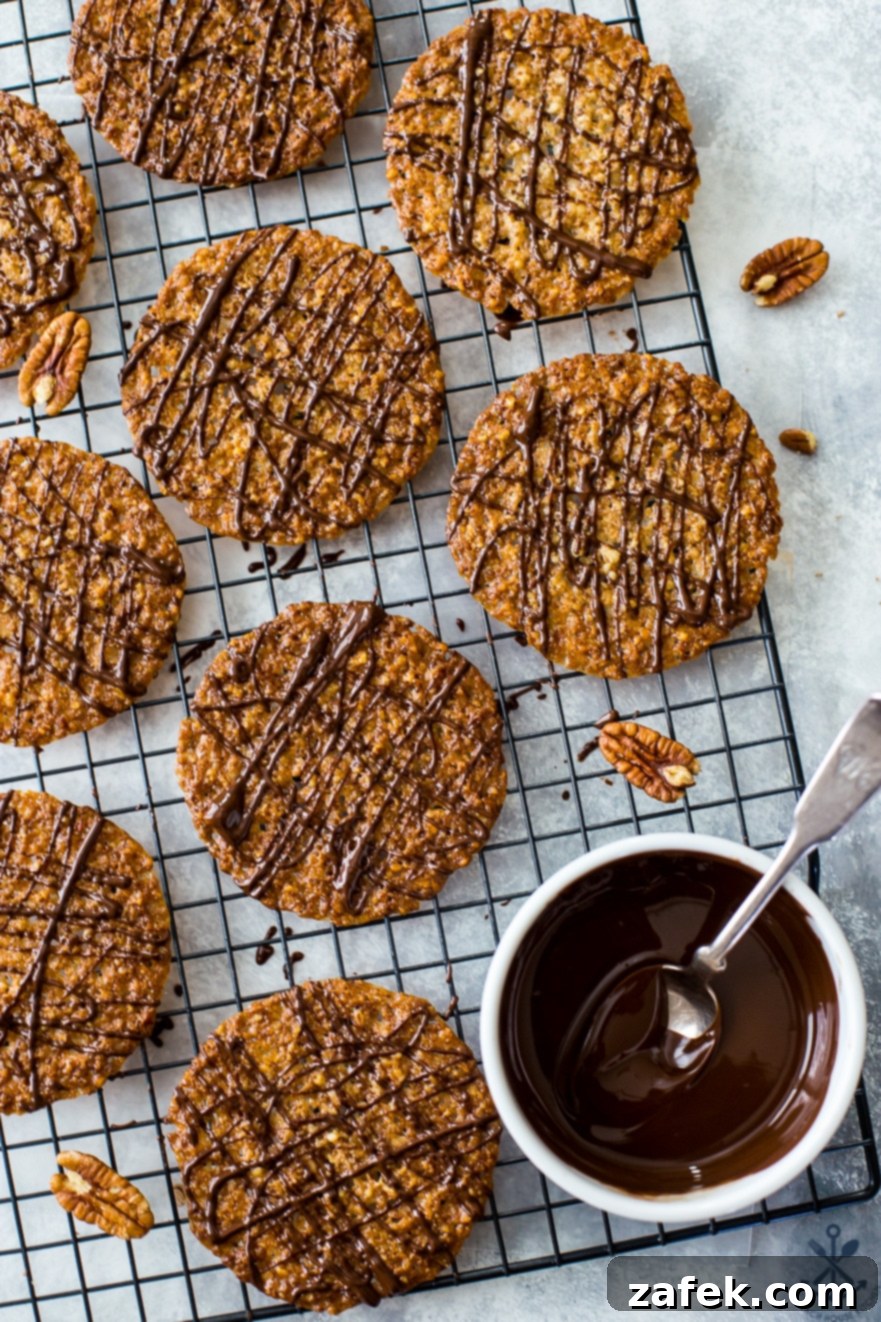 Overhead photo of delicate lace cookies with chocolate drizzle on a wire rack, next to a small bowl of melted chocolate