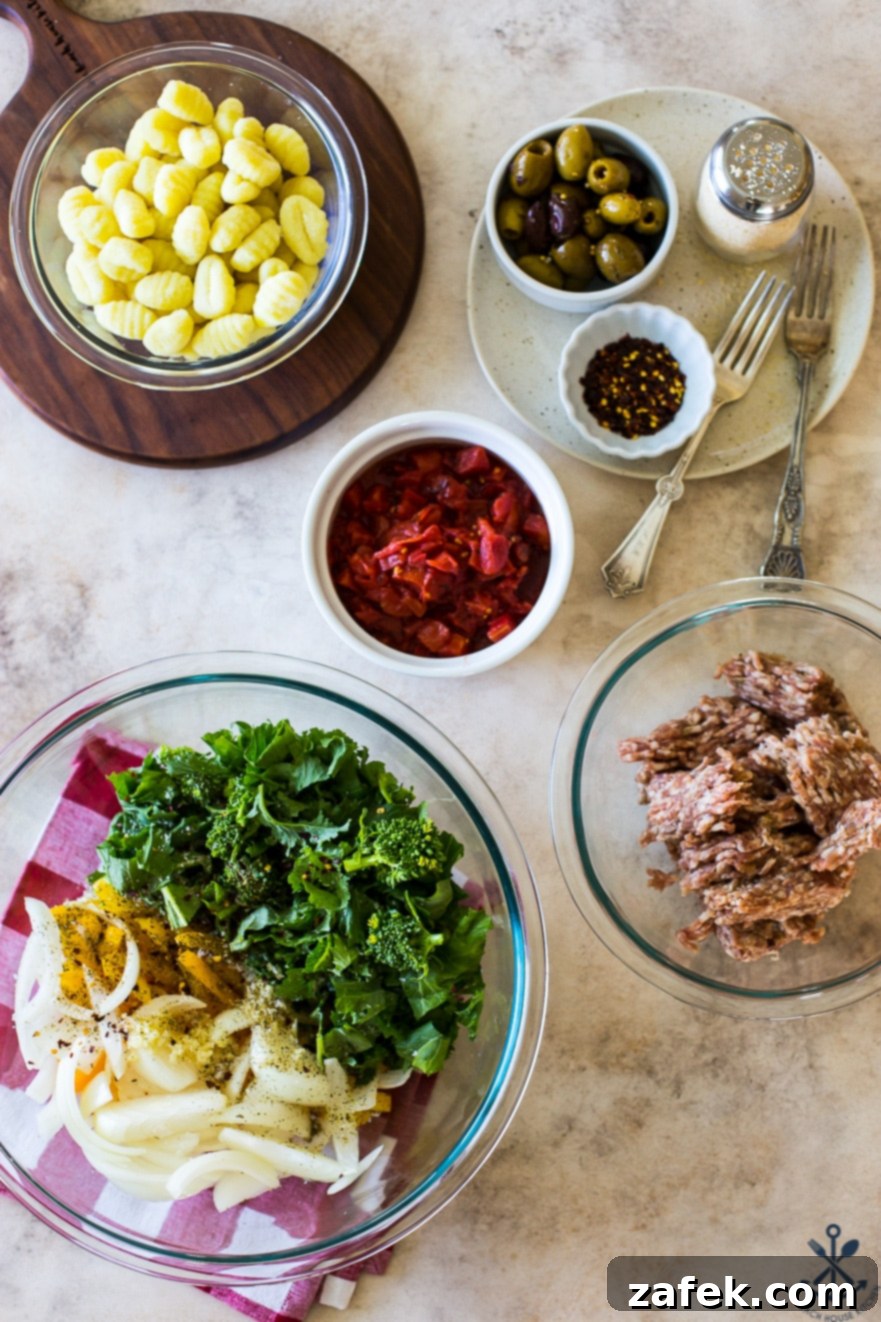 Overhead photo of individual bowls filled with prepped ingredients, including chopped broccoli rabe, sliced bell peppers, onions, diced tomatoes, gnocchi, and bulk Italian sausage, ready for the sheet pan.