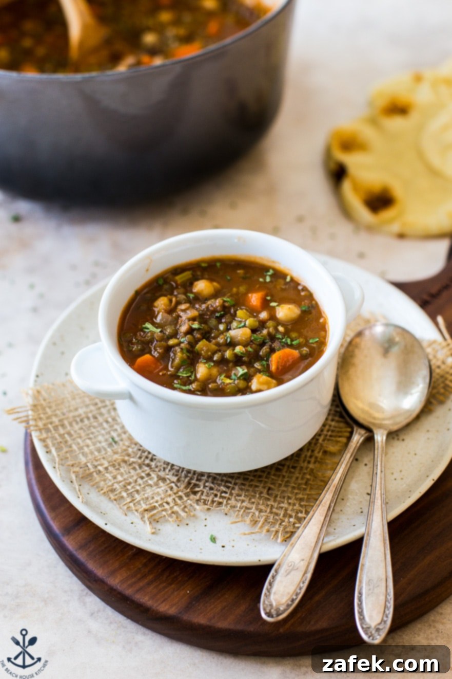 Comforting Lentil and Chickpea Soup 7 Up close photo of a white bowl filled with steaming lentil and chickpea soup, placed on a plate with two spoons