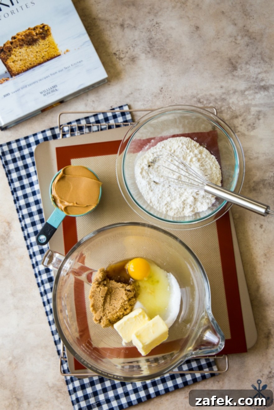 Overhead photo of ingredients for peanut butter cookies in glass bowls on a silpat lined tray with a blue and white checked napkin