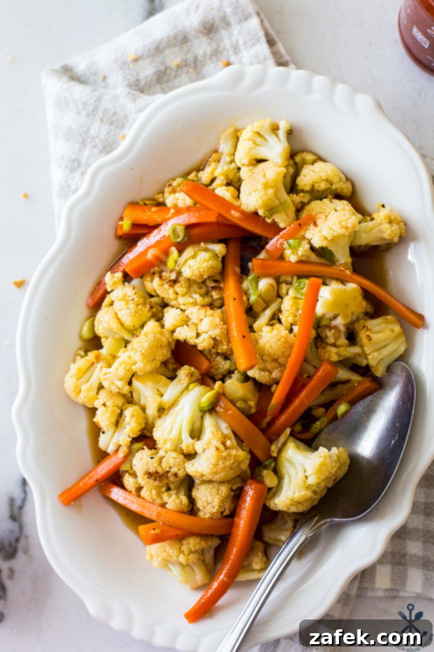 Up close overhead photo of roasted cauliflower and carrots on a white oval dish with a spoon, showing the glossy sauce