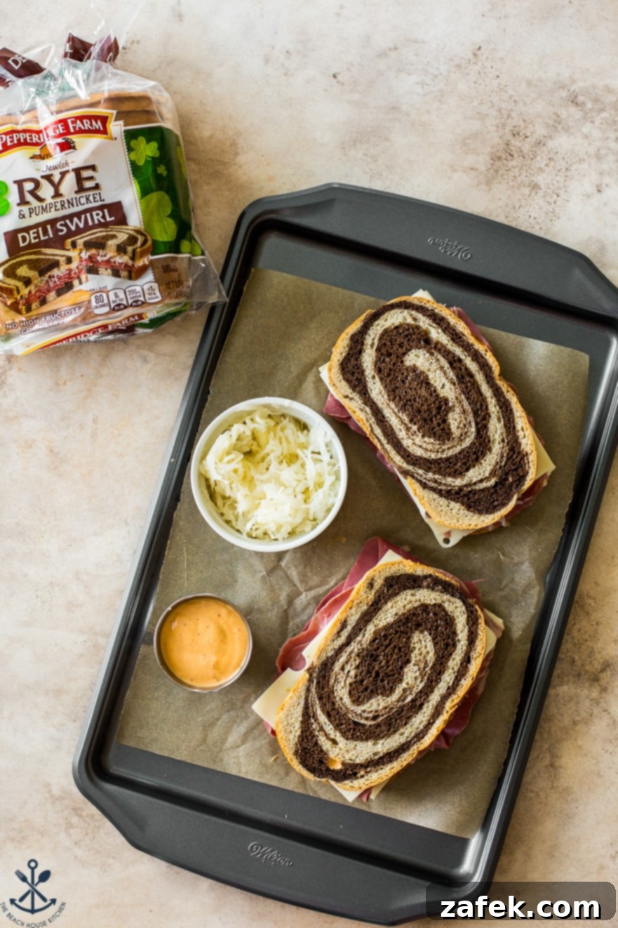 An overhead view of two assembled Reuben sandwiches, ready for grilling, placed on a baking sheet. Beside them are bowls of sauerkraut and Russian dressing, highlighting the key ingredients.