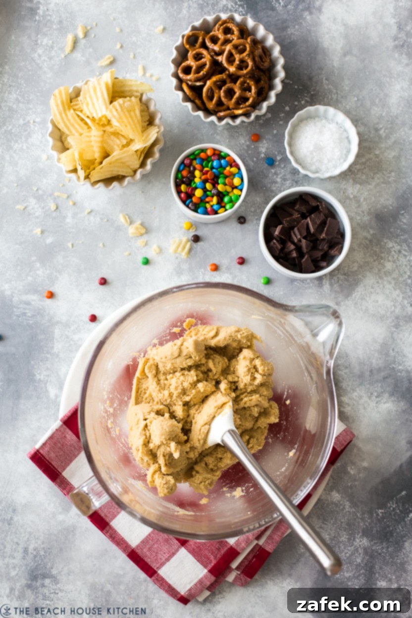Kitchen Sink Cookies 4 Overhead photo showcasing the array of key ingredients for kitchen sink cookies: flour, sugars, eggs, butter, M&M's, pretzels, chocolate, and potato chips