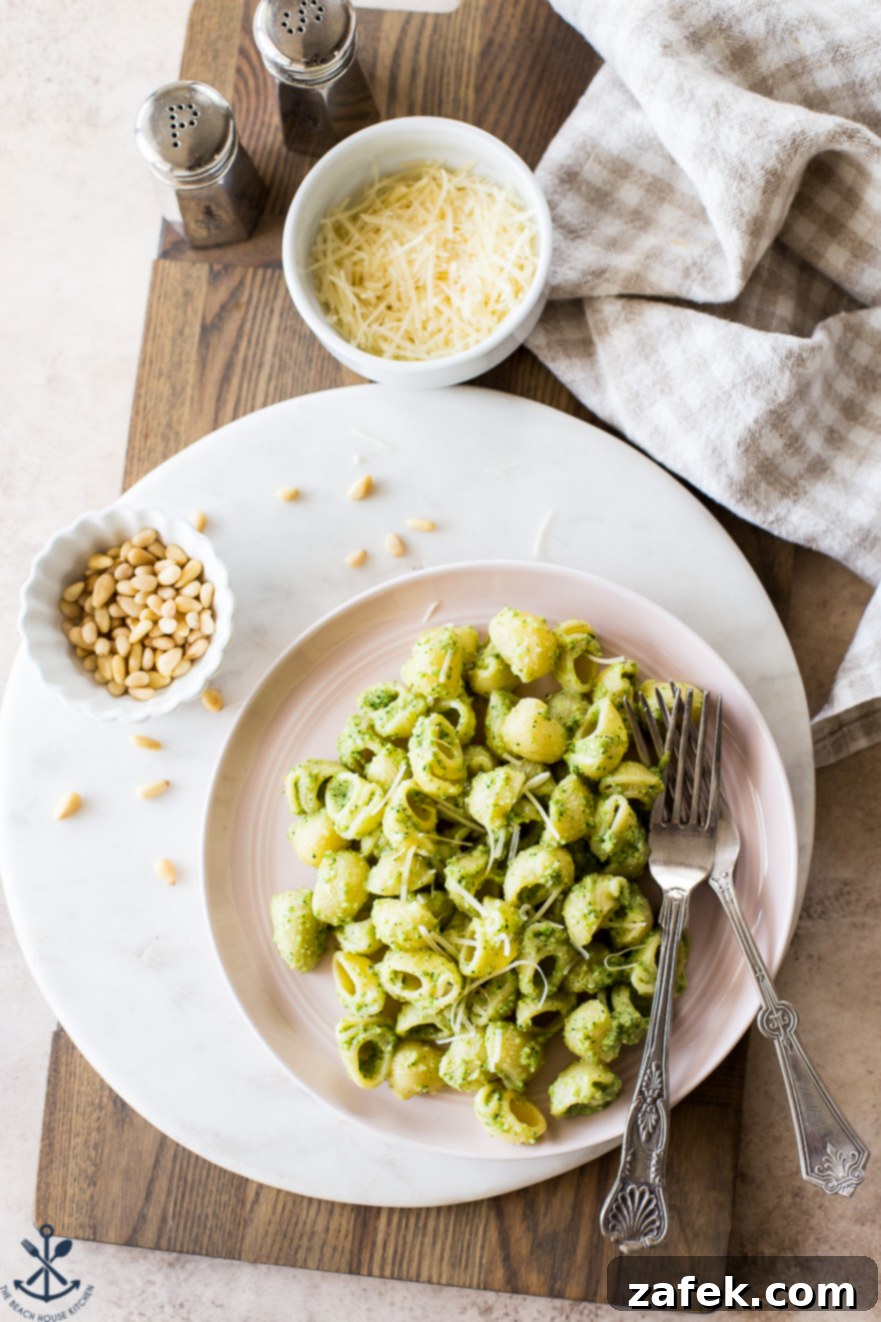 Overhead photo of a pink plate of Broccoli Pesto Pasta on a round marble board with a small bowl of pine nuts, ready to be served