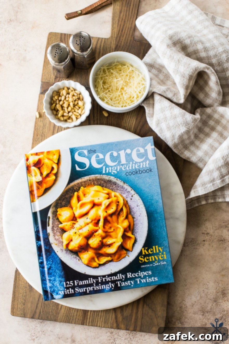 Overhead photo of The Secret Ingredient Cookbook open on a round marble board, accompanied by a small bowl of pine nuts and shredded Parmesan cheese