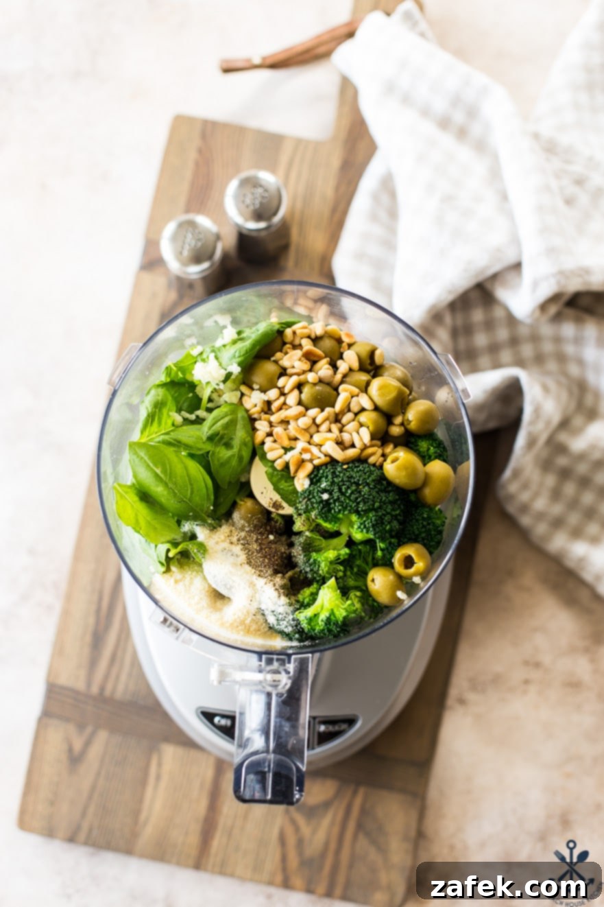Overhead photo of a food processor filled with fresh broccoli, green olives, pine nuts, and basil, ready to be blended into pesto