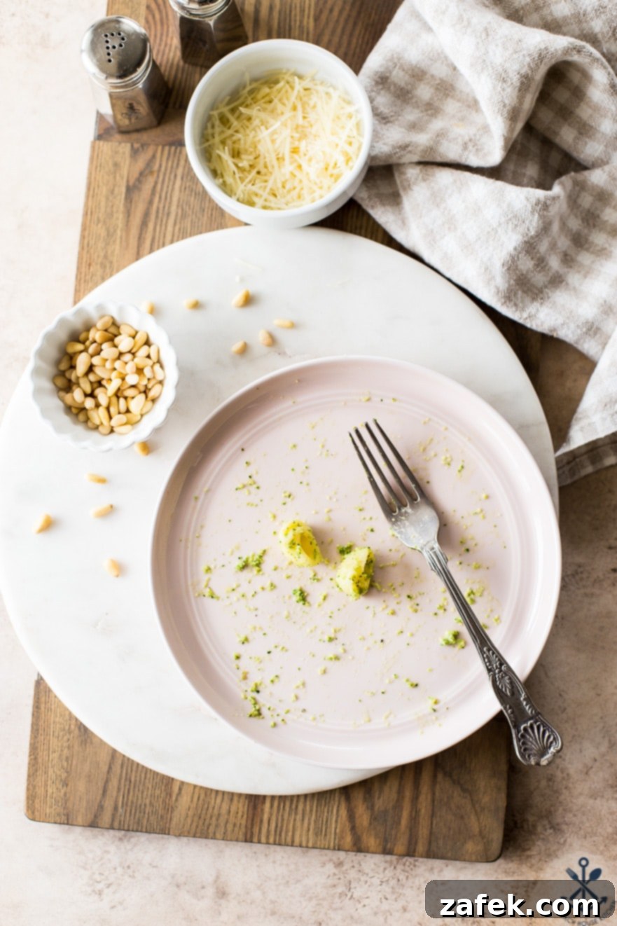 Overhead photo of a light pink plate with two large shell noodles coated in pesto and a fork, with a small bowl of pine nuts off to the side