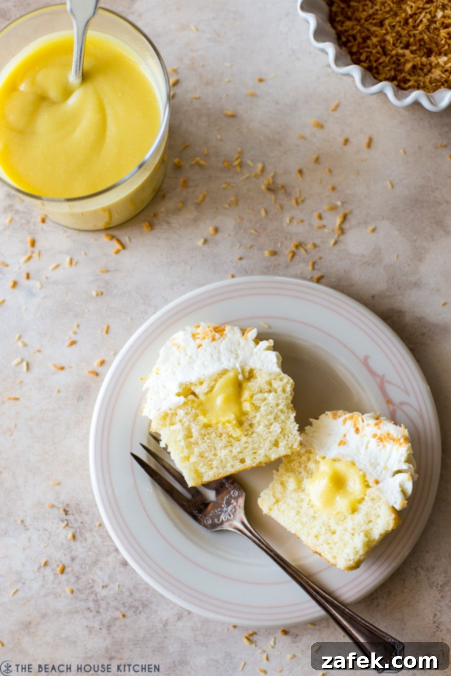 Overhead close-up photo of a single Piña Colada Cupcake, cut open to reveal the bright pineapple curd filling, placed on a white plate with a fork and a small glass of curd.