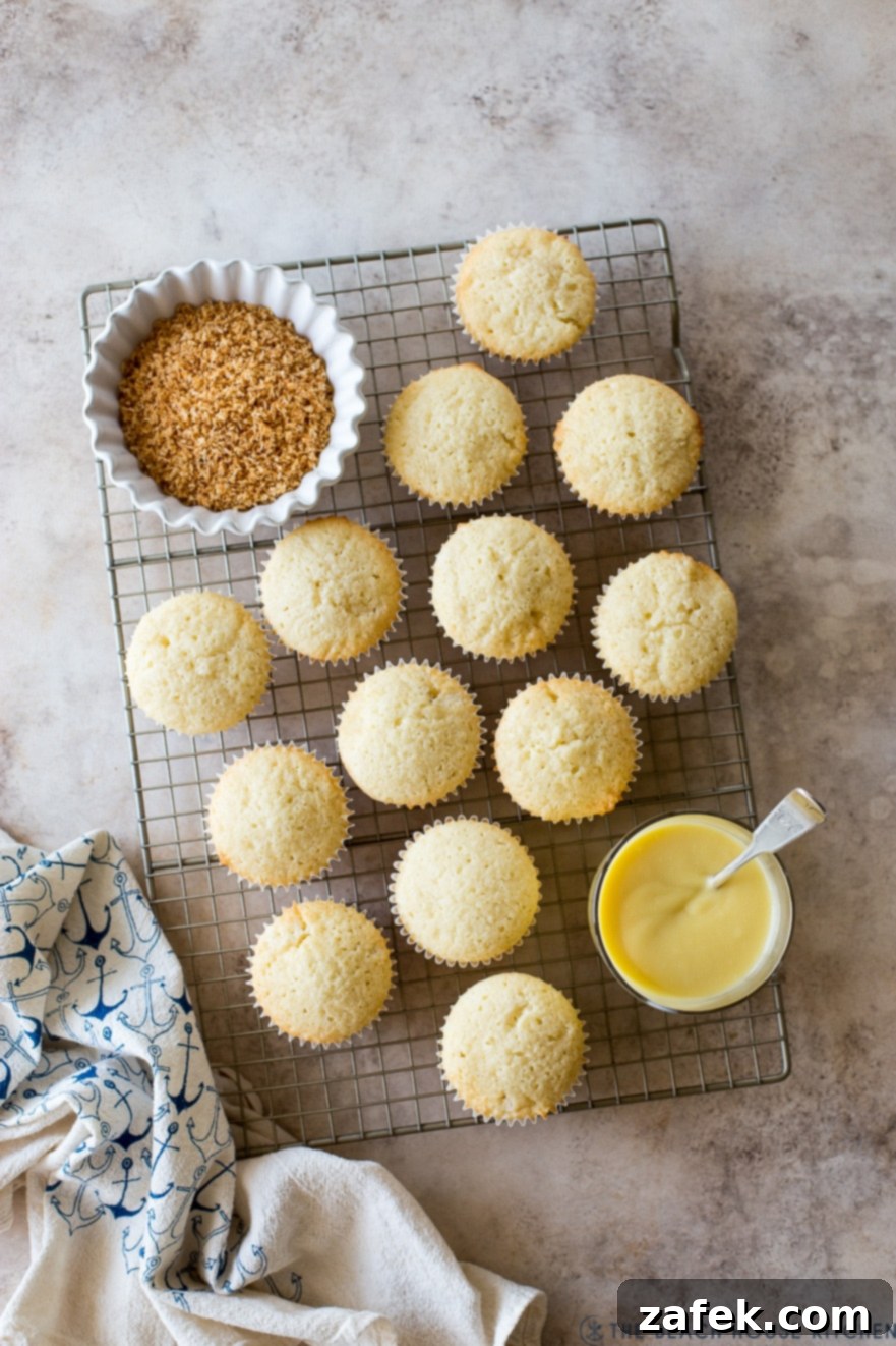 Overhead view of several unfrosted coconut cupcakes cooling on a wire rack, with a jar of homemade pineapple curd and a bowl of golden toasted coconut flakes nearby.