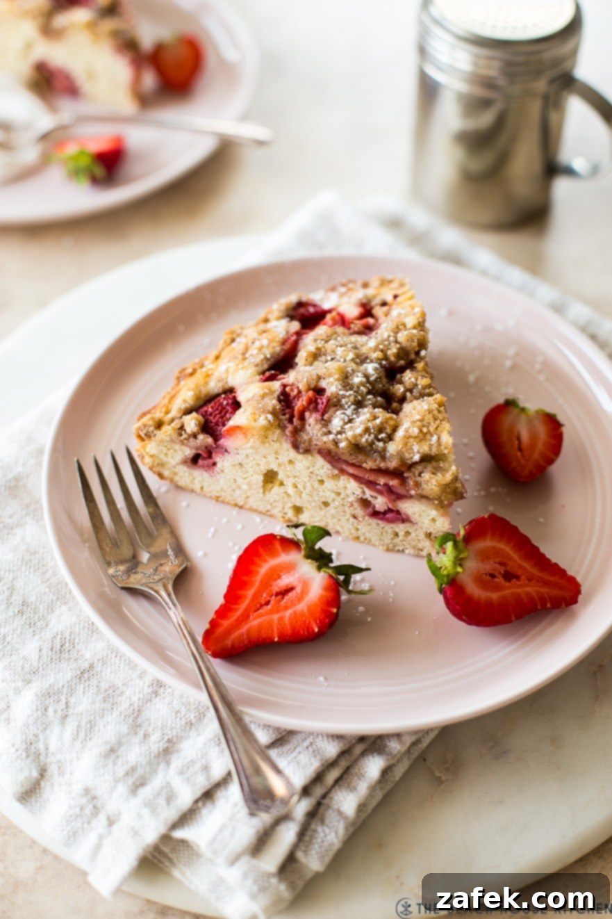 Sweet Strawberry Streusel 3 A perfect slice of warm Strawberry Buckle, showcasing its moist cake, juicy baked strawberries, and crunchy streusel, resting on a pink plate with fresh strawberry slices and a fork.