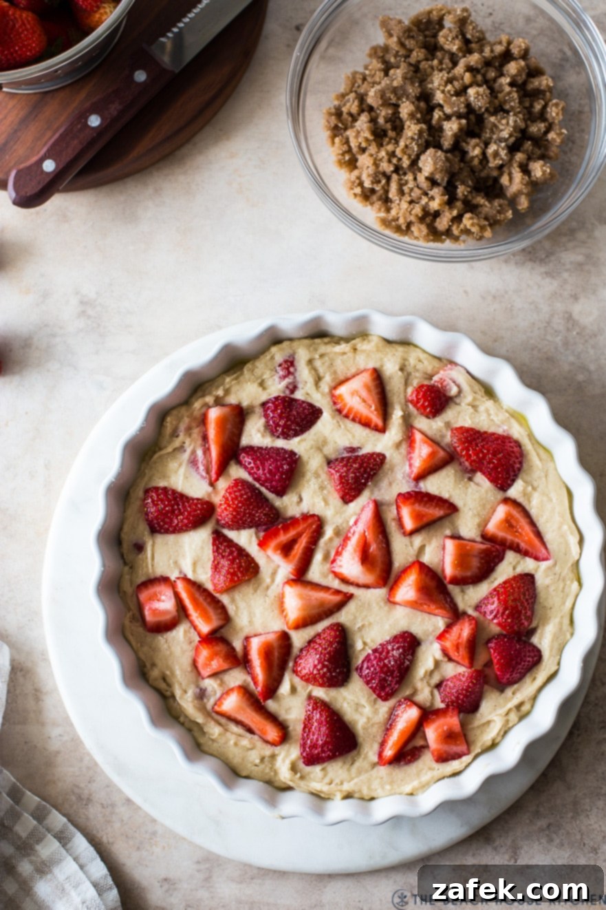 Sweet Strawberry Streusel 4 An unbaked Strawberry Buckle, featuring a smooth cake batter topped generously with fresh halved strawberries and a crumbly streusel mixture, prepared in a white baking dish before going into the oven.