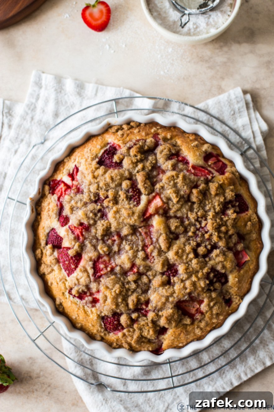 Sweet Strawberry Streusel 6 Close-up detail of a freshly baked Strawberry Buckle in a white round baking dish, its golden-brown streusel and tender cake visible, presented on a rustic tan and white checked napkin.