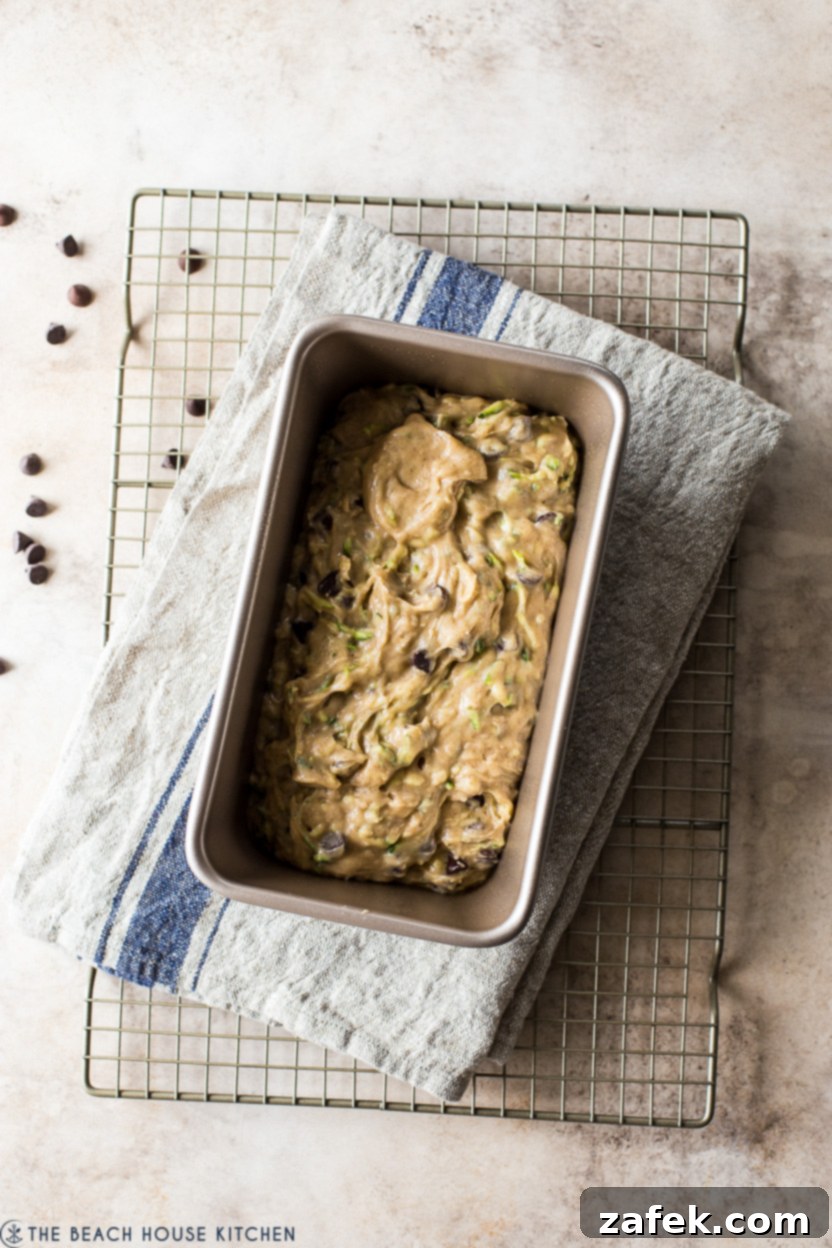 Decadent Chocolate Chip Zucchini Loaf 9 Overhead photo of unbaked zucchini bread batter in a loaf pan, topped with chocolate chips, sitting on a wire rack before going into the oven.