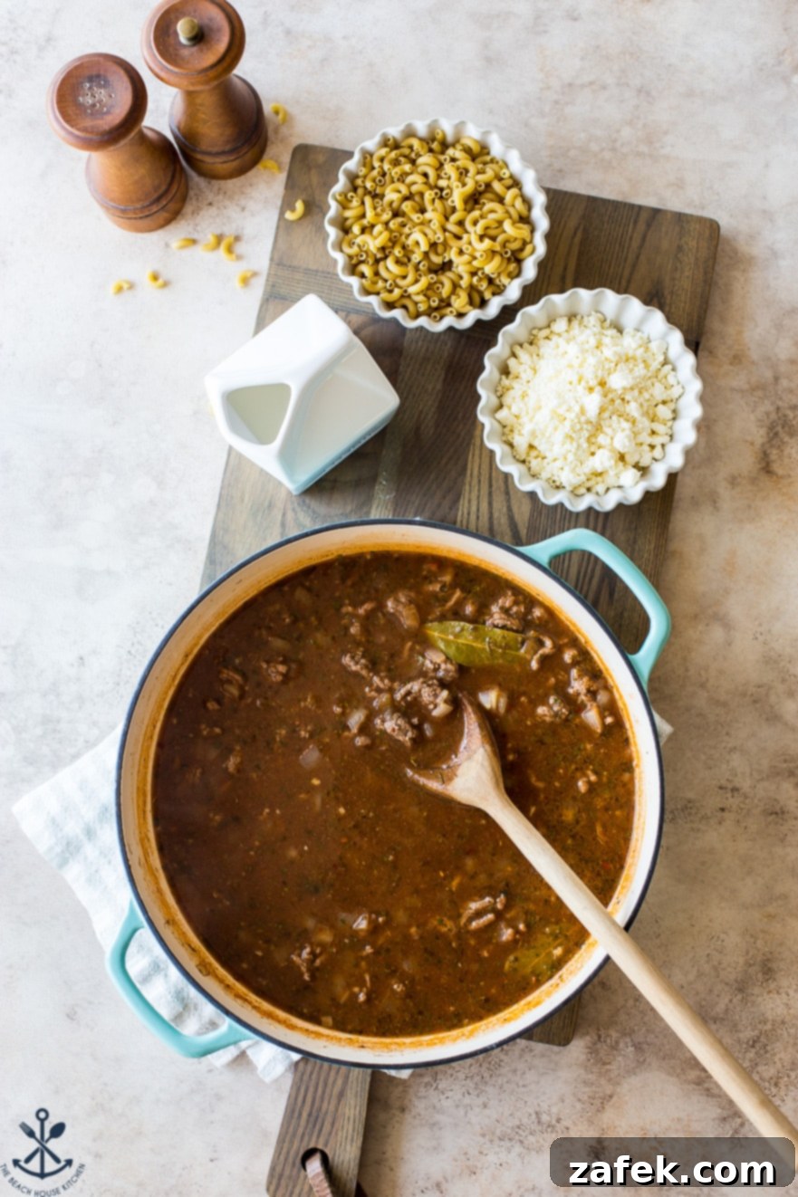 Homemade Greek Beef and Orzo Skillet 7 Overhead photo of a skillet filled with beef and broth and a bay leaf on a wooden board with a bowl of dried pasta, a bowl of feta crumbles and a small container of milk