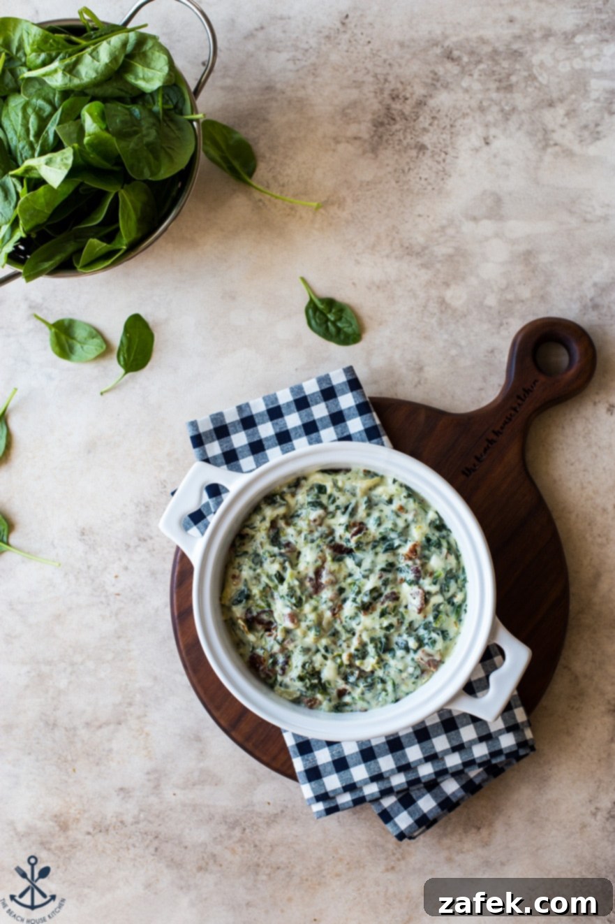 Ultimate Bacon Spinach Artichoke Dip 3 Overhead photo of creamy, unbaked spinach artichoke dip mixture in a white baking dish on a rustic wooden board, with fresh spinach leaves in a small bowl on the side