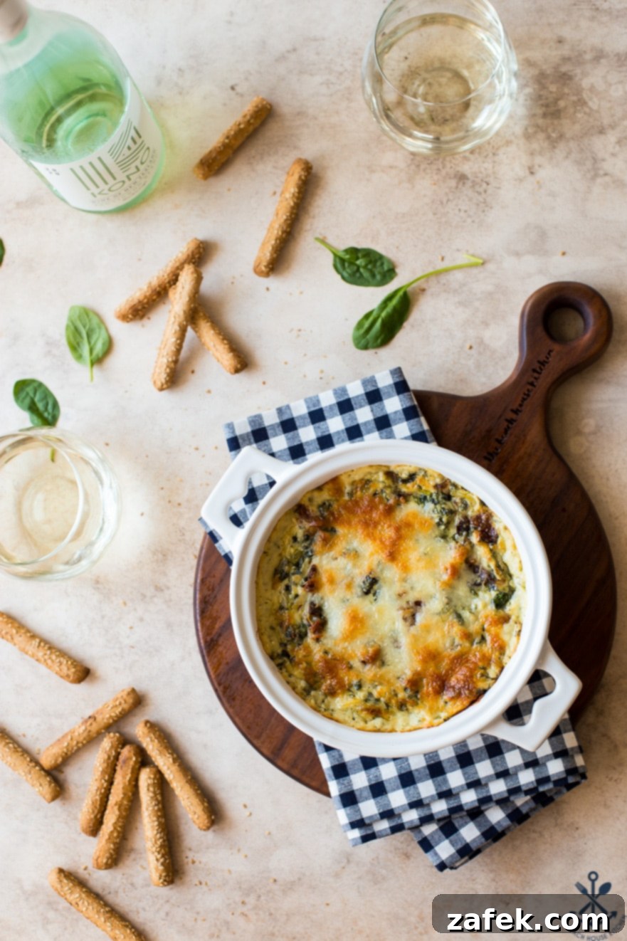 Ultimate Bacon Spinach Artichoke Dip 6 Overhead photo of a white baking dish filled with spinach artichoke dip, surrounded by toasted breadsticks and two wine glasses, creating a cozy appetizer scene