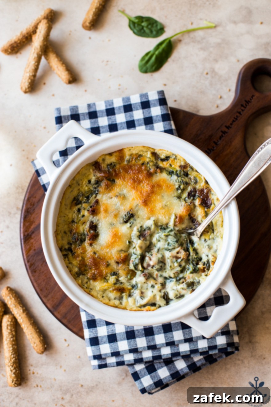 Ultimate Bacon Spinach Artichoke Dip 7 Overhead photo of a serving spoon partially submerged in a warm, cheesy spinach artichoke dip in a white baking dish
