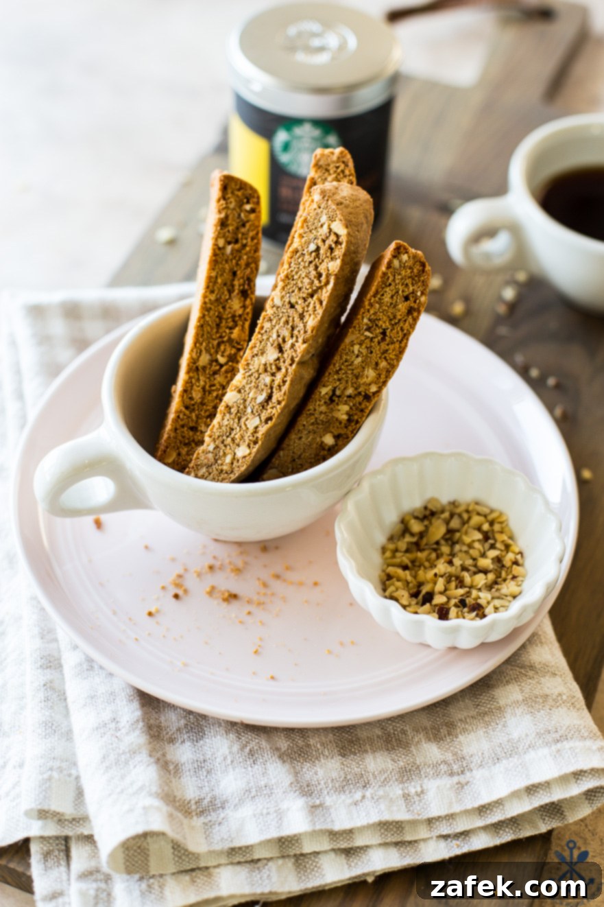 Slices of plain hazelnut biscotti neatly arranged in a coffee cup, with a small dish of raw hazelnuts on a pink plate, before being dipped in chocolate