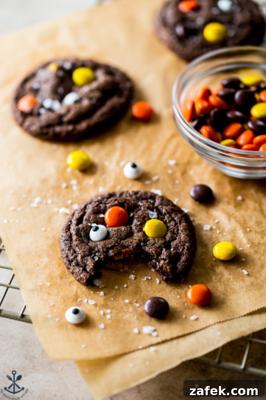 A dark chocolate cookie on parchment paper, adorned with colorful Reese's Pieces and whimsical googly eyes, with a blurred background of candy
