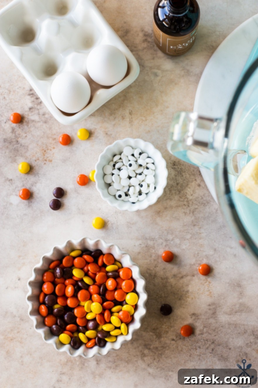 Overhead shot showcasing a bowl of colorful Reese's Pieces, a small bowl of edible googly eyes, and a bottle of vanilla extract, hinting at the baking magic to come
