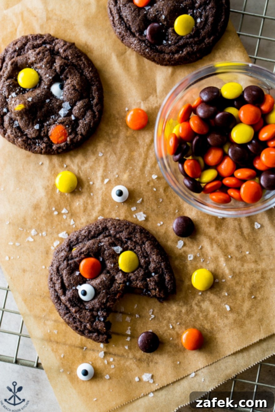 An overhead view of freshly baked chocolate cookies, generously adorned with Reese's Pieces and playful googly eyes, next to a bowl of extra Reese's candy