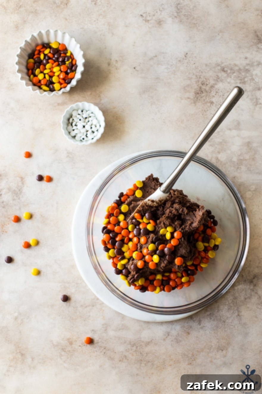 An overhead view of raw chocolate cookie dough, generously mixed with Reese's Pieces, alongside a small bowl of extra candies and another bowl of googly eyes, ready for assembly