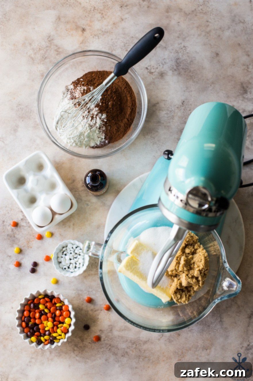 A vibrant turquoise stand mixer bowl containing brown sugar and butter, surrounded by fresh eggs, bowls of Reese's Pieces and googly eyes, and a bowl of sifted flour and cocoa with a whisk