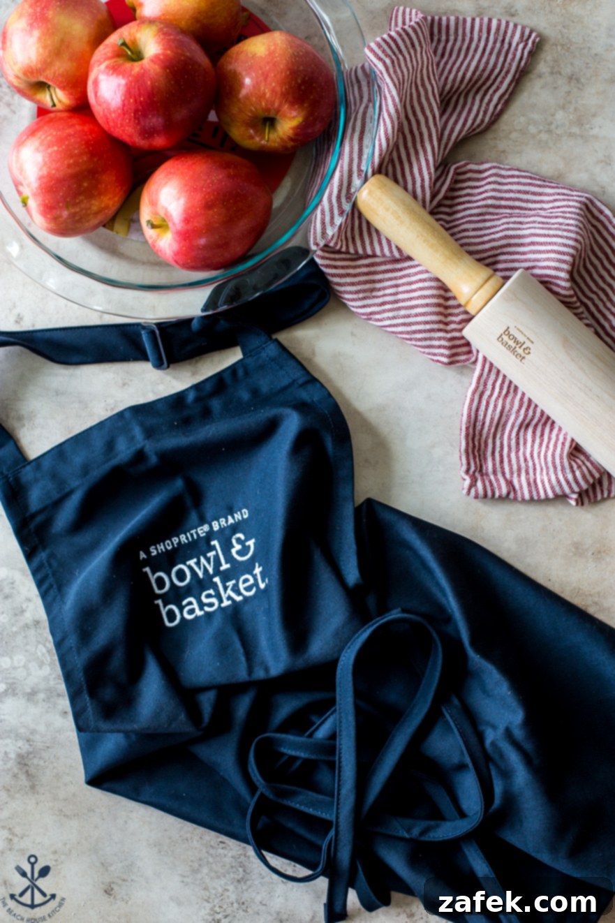 ShopRite's Holiday Apple Pie Fest 4 Overhead photo featuring a navy blue Bowl & Basket apron, a pie plate brimming with fresh apples, and a wooden rolling pin, highlighting the preparation for the ShopRite apple pie.