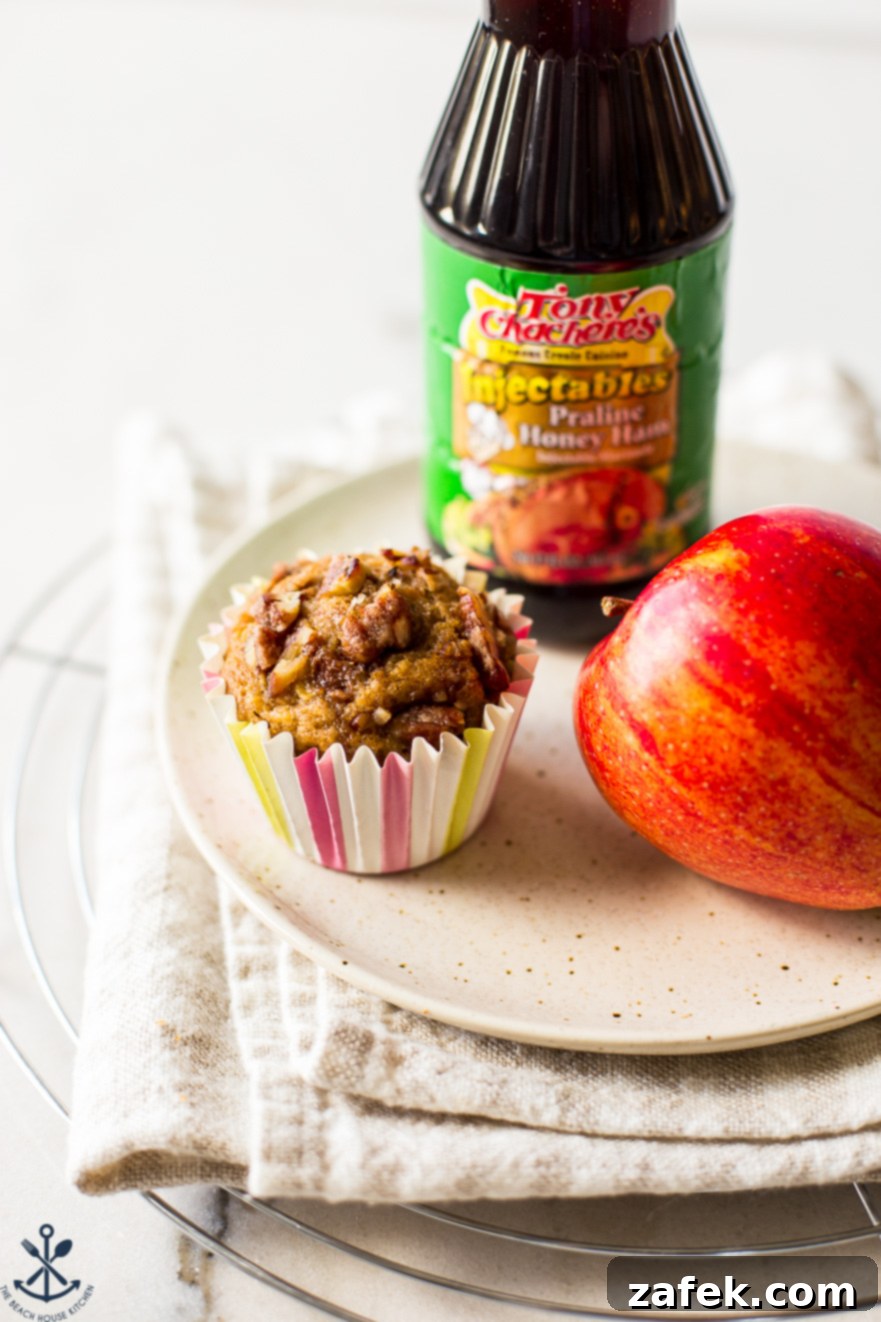 Golden Apple Praline Muffins 2 A beautifully baked Apple Praline Muffin on a pristine white plate, showcasing its golden-brown exterior and enticing candied pecan topping. In the background, a fresh red apple and a bottle of Tony Chachere's Praline Honey Ham Marinade hint at the key ingredients, inviting viewers to savor its unique flavor.