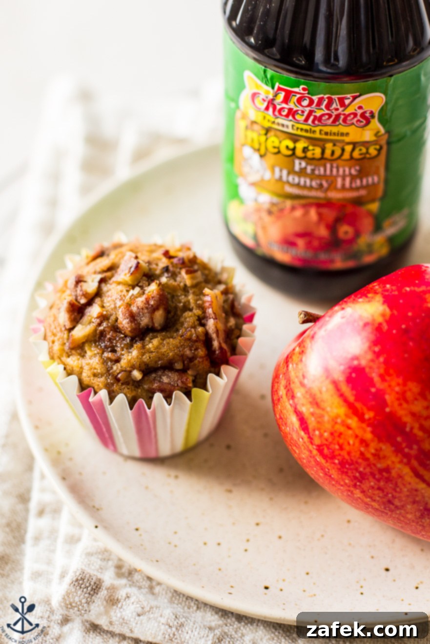 Golden Apple Praline Muffins 6 A single, perfectly baked Apple Praline Muffin takes center stage on a white plate, its golden crust and visible pecan pieces invitingly displayed. A fresh apple and the distinctive bottle of praline honey ham marinade provide a colorful backdrop, underscoring the muffin's key components.