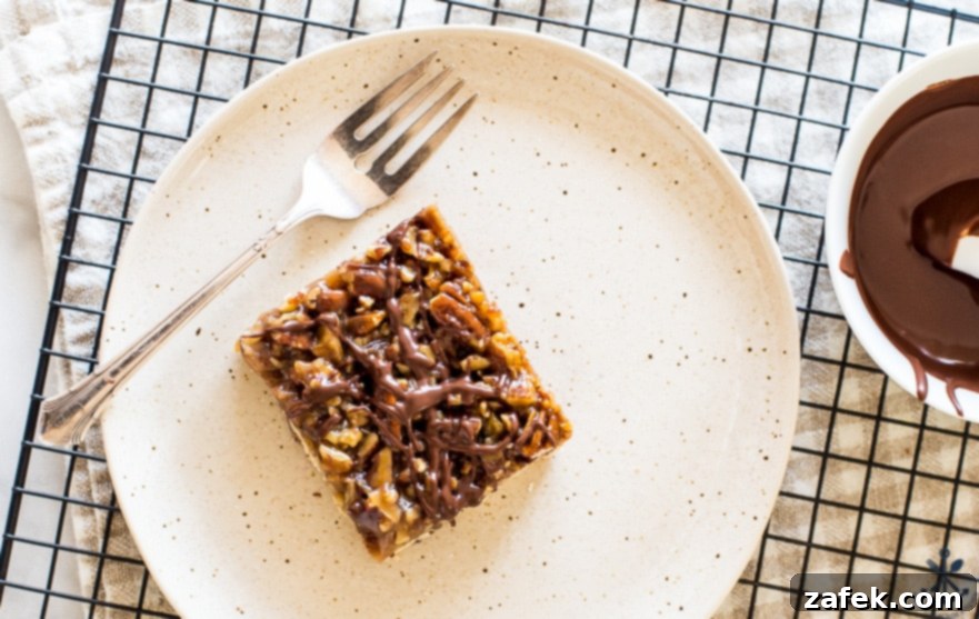 Decadent Maple Pecan Bars Adorned with Chocolate 6 Close-up overhead photo of a single chocolate drizzled maple pecan bar on a white plate with a fork, highlighting the glossy chocolate and nutty topping