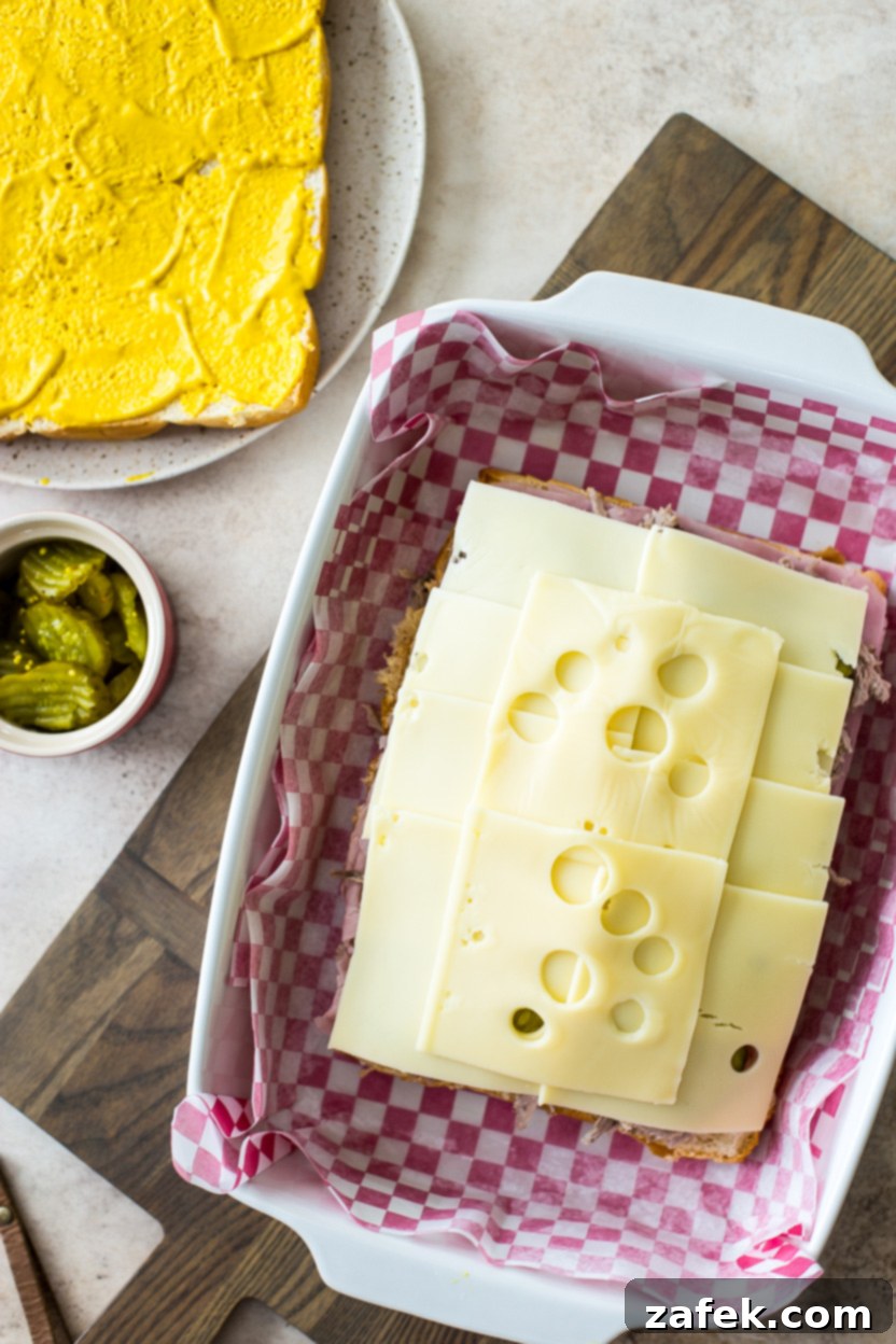 Savory Cuban Mini Sandwiches 5 Overhead photo of Swiss cheese topped sliders in a baking dish with bread topped with yellow mustard off to the side and a small bowl of pickles.
