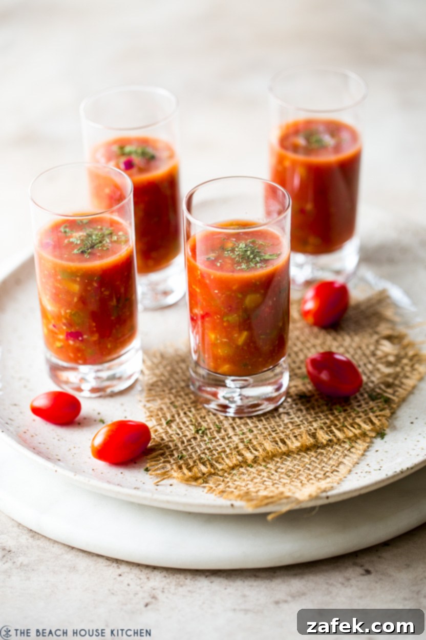 Four shooters glasses filled with vibrant red gazpacho, garnished with herbs, on a round marble board.