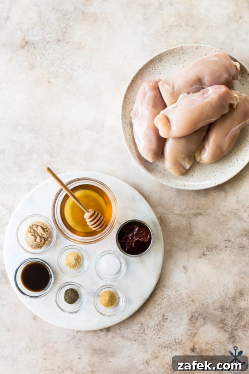 Sticky Slow Cooker Honey Chipotle Chicken 3 Overhead photo of uncooked boneless, skinless chicken breasts arranged on a round marble board, surrounded by bowls of honey, chipotle peppers in adobo sauce, brown sugar, and seasonings for the sauce