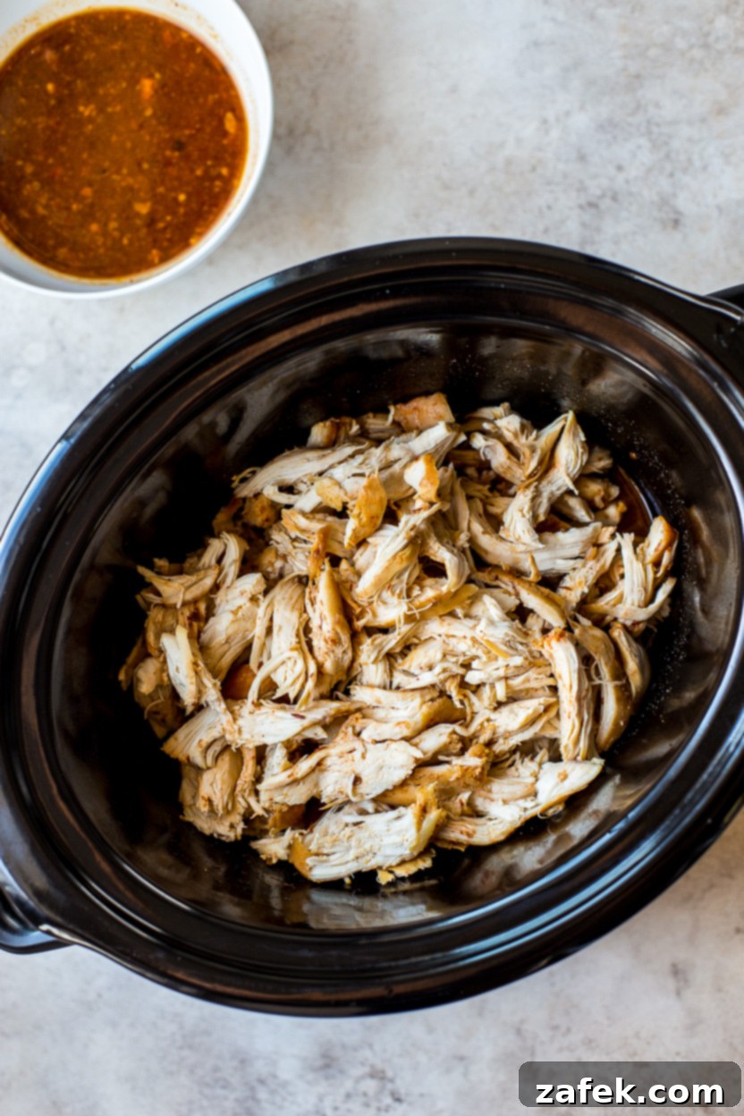 Sticky Slow Cooker Honey Chipotle Chicken 7 Overhead photo of shredded honey chipotle chicken mixed with thickened sauce in a slow cooker, with a small bowl of fresh sauce on the side for garnish