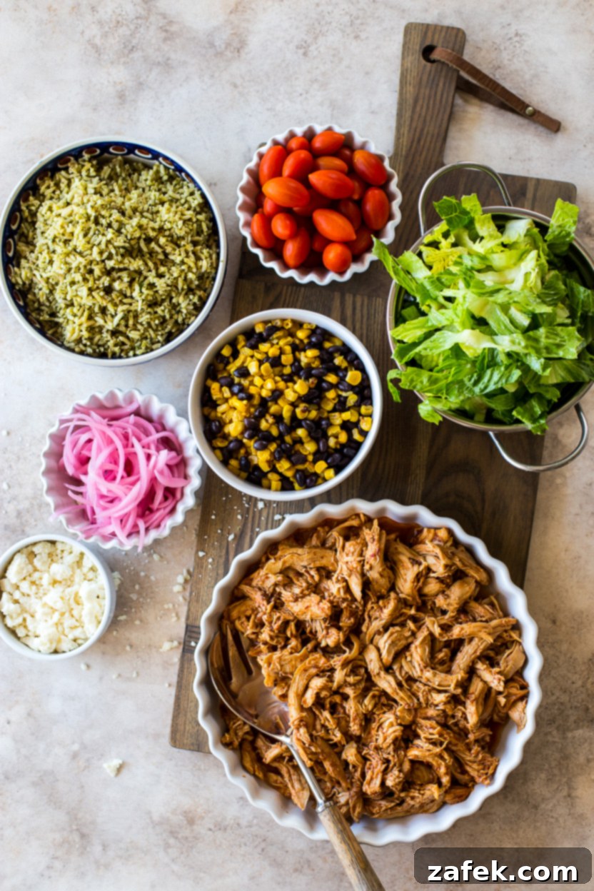 A top-down shot of various ingredients for Honey Chipotle Chicken Bowls, including cooked chicken, green rice, pickled onions, corn, black beans, tomatoes, and Cotija cheese.