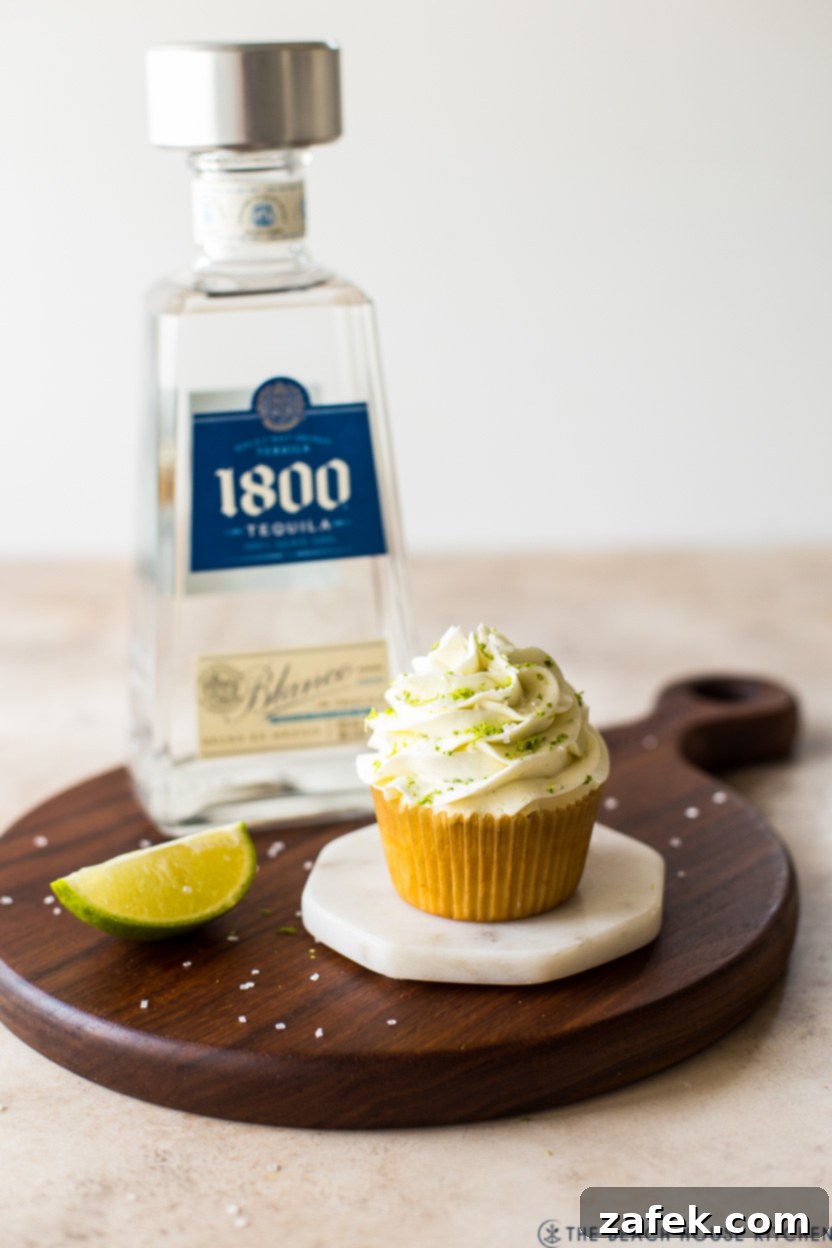 A single, perfectly frosted margarita cupcake resting on a white marble coaster, with a blurred bottle of tequila in the background