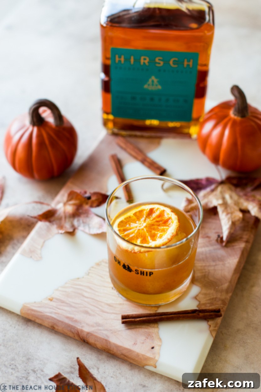 Fireside Pumpkin Old Fashioned 2 A Pumpkin Spice Old Fashioned cocktail on a rustic board surrounded by bourbon, pumpkins, fall leaves, and cinnamon sticks, evoking autumn warmth.