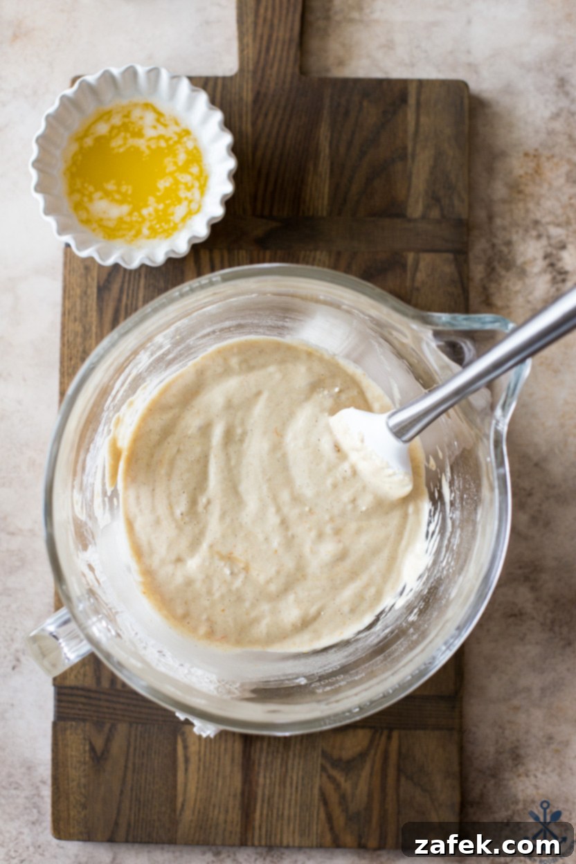 Overhead photo of a clear glass bowl containing madeleine batter, with a small bowl of melted butter next to it.