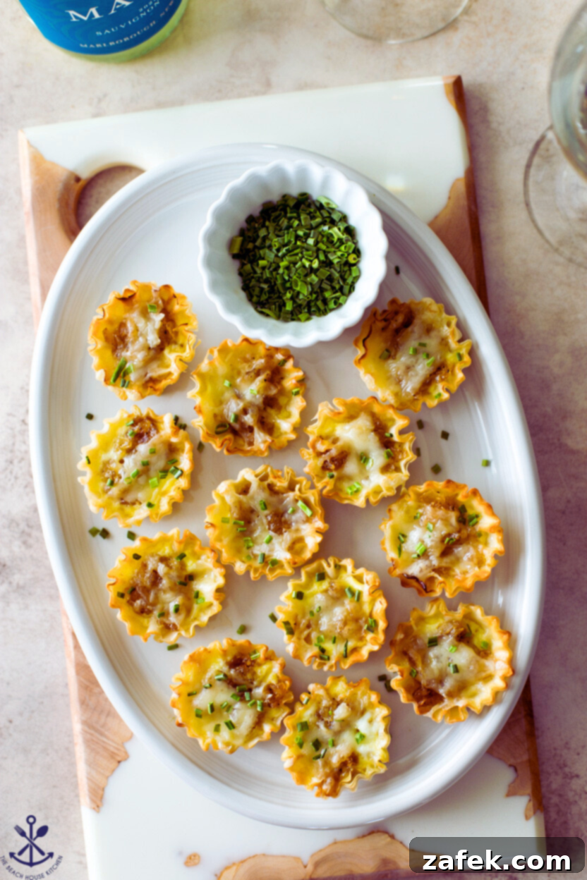 Overhead photo of French onion soup bites on an oval platter with a small bowl of chopped chives, highlighting their golden brown crust and flaky pastry