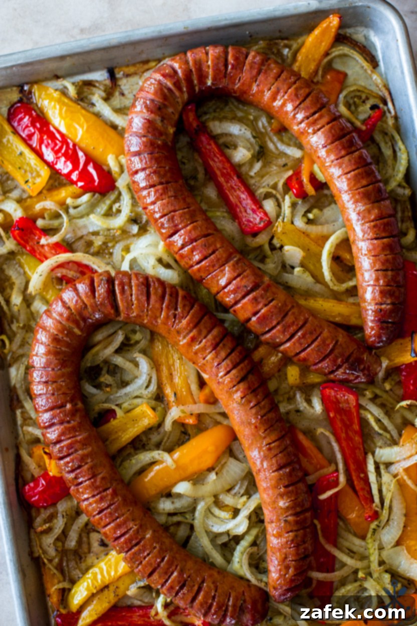 An inviting overhead photograph showcasing two rings of beautifully scored Hasselback kielbasa, perfectly nestled on a bed of seasoned onions and bell peppers on a large sheet pan.