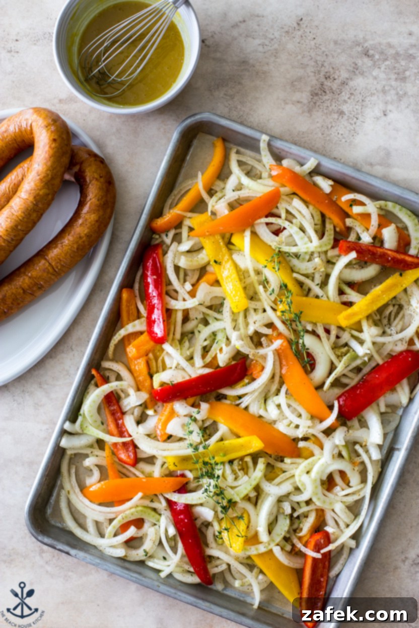 A spacious sheet pan filled with seasoned onions and bell peppers, ready to be placed in the oven, with two rings of Hasselback kielbasa awaiting their turn on the side.