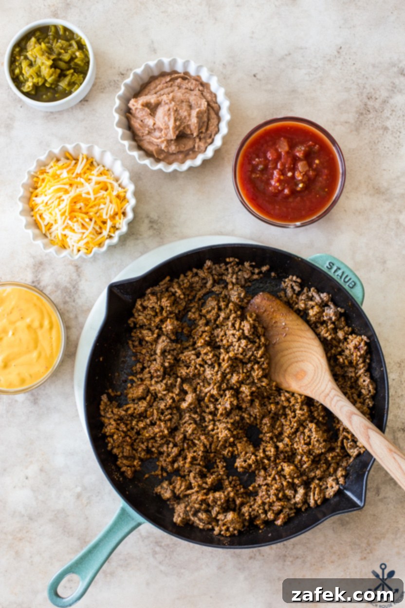 Overhead photo of a skillet containing cooked, perfectly seasoned taco meat, surrounded by small bowls of shredded cheddar jack cheese, vibrant red salsa, creamy refried beans, and bright diced green chiles, all ready for assembly