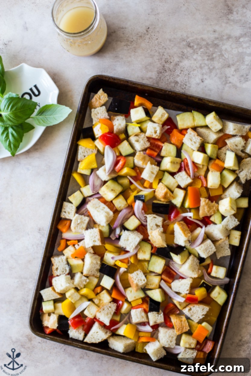 Overhead photo of a pre-baked Sheet Pan Roasted Vegetable Panzanella with uncooked bread cubes, eggplant, bell peppers, and shallots on a baking sheet, seasoned and ready for the oven.