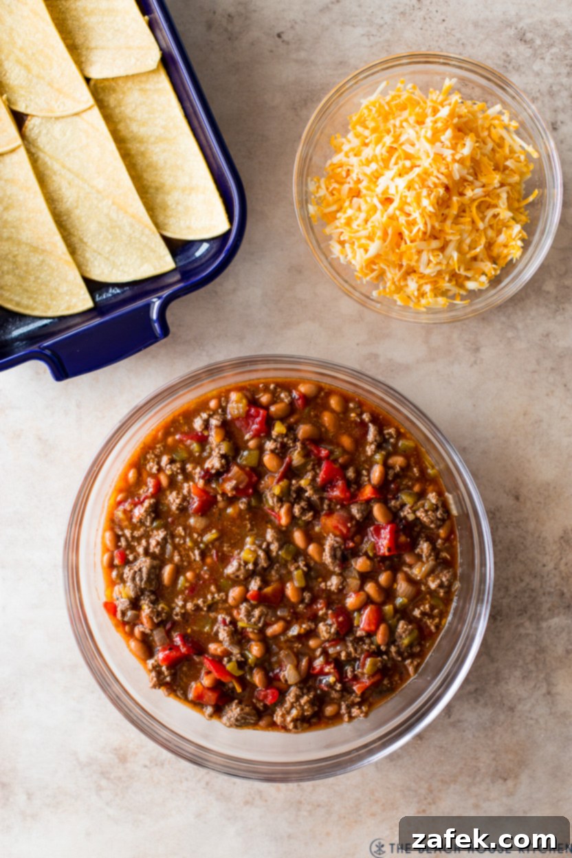 Overhead photo of a meat mixture in a bowl with green chiles, pinto beans, and tomatoes, alongside a bowl of shredded cheddar jack cheese